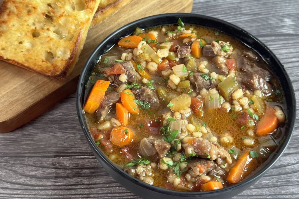 Beef and barley soup in a black bowl with tender beef chunks, pearl barley, carrots, and vegetables in a brothy tomato-beef base, served with toasted bread.