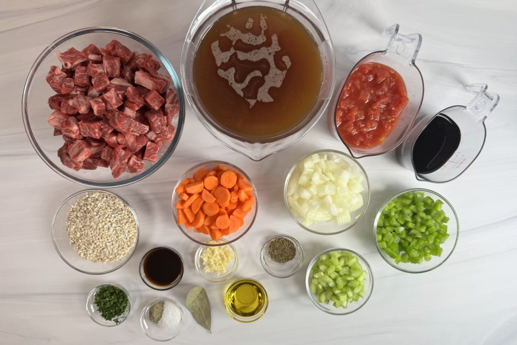 Ingredients for beef and barley soup including beef chuck, pearl barley, carrots, celery, onion, beef broth, diced tomatoes, garlic, thyme, bay leaf, Worcestershire sauce, and olive oil.