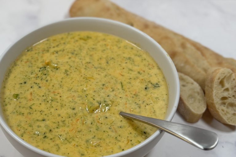 Creamy broccoli cheddar soup in a white bowl with a spoon, served with sliced baguette on a marble surface.