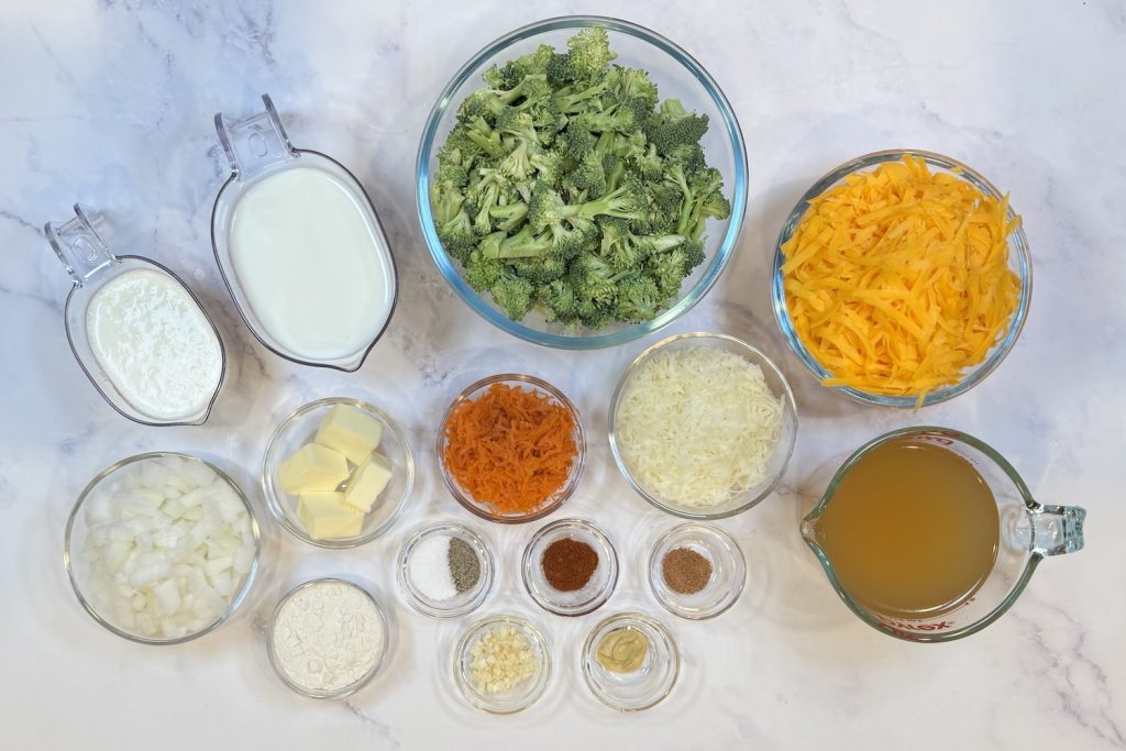 Ingredients for broccoli cheddar soup including fresh broccoli florets, shredded cheddar and Gruyère, milk, half-and-half, broth, diced onion, butter, flour, grated carrot, garlic, Dijon mustard, and seasonings arranged in glass bowls.