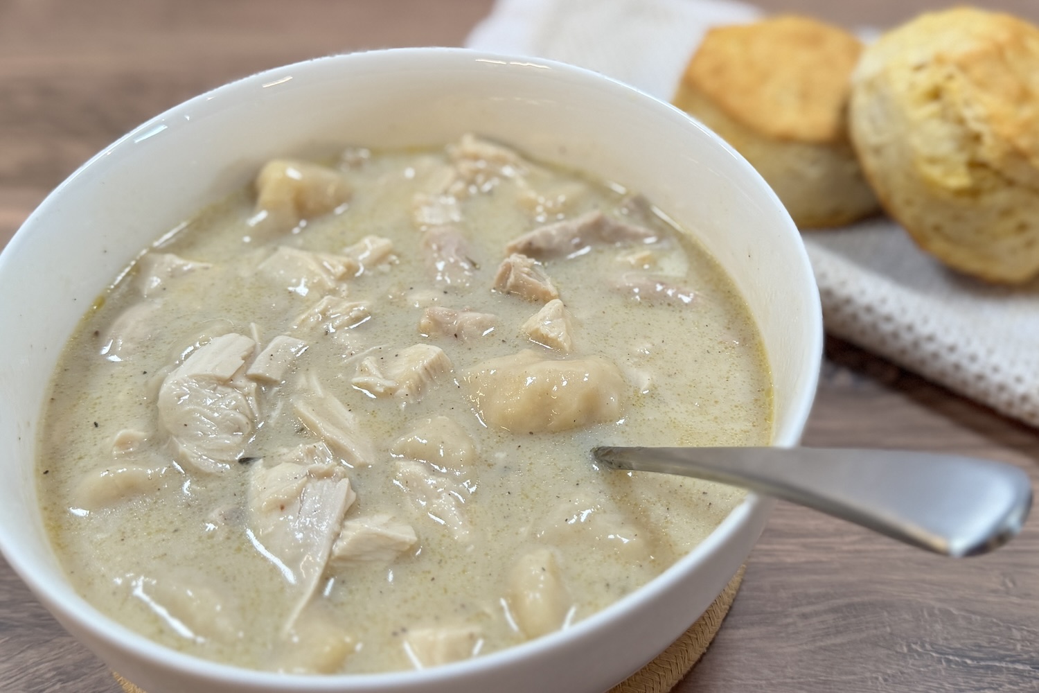 Creamy chicken and dumplings soup in a white bowl with tender chicken pieces and soft dumplings, served with golden biscuits on a wooden surface.