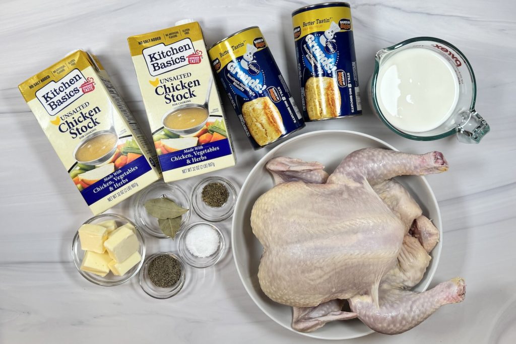 Ingredients for chicken and dumplings soup including whole raw chicken, chicken stock, refrigerated biscuit dough, milk, butter, bay leaves, thyme, salt, and pepper arranged on a marble surface.