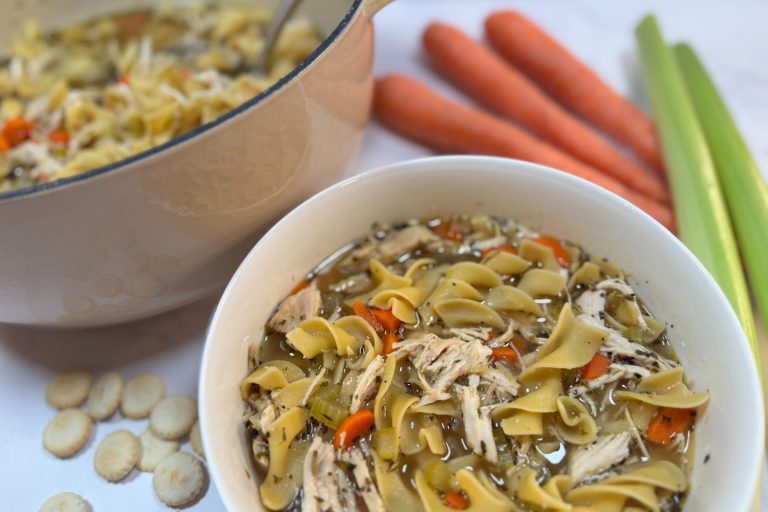 Bowl of homemade chicken noodle soup with shredded chicken, egg noodles, carrots, and celery, with Dutch oven in background.