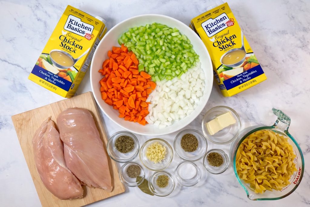 Ingredients for chicken noodle soup including chicken breasts, carrots, celery, onion, egg noodles, butter, garlic, dried herbs, and chicken stock.