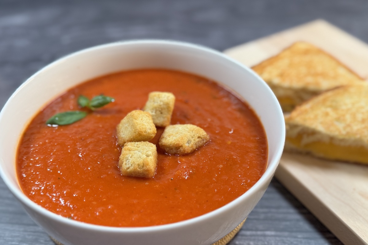 Creamy tomato soup topped with croutons and basil in a white bowl, Panera copycat recipe.