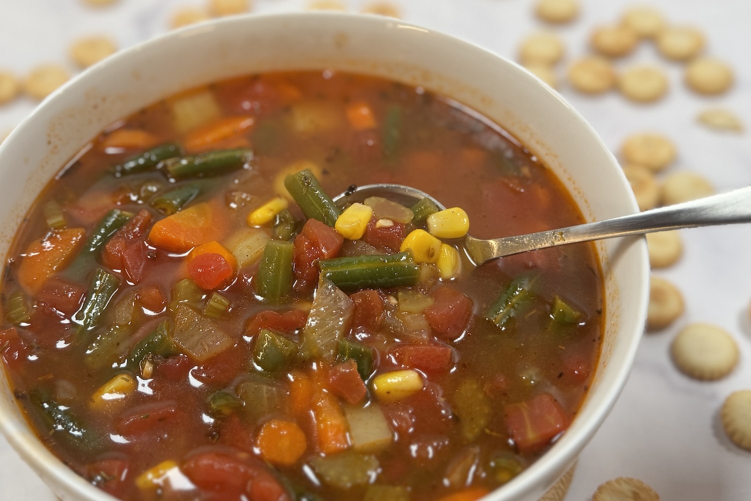 Close-up of brothy vegetable soup in a white bowl with carrots, green beans, corn, potatoes, and diced tomatoes, with a spoon lifting a scoop from the bowl.