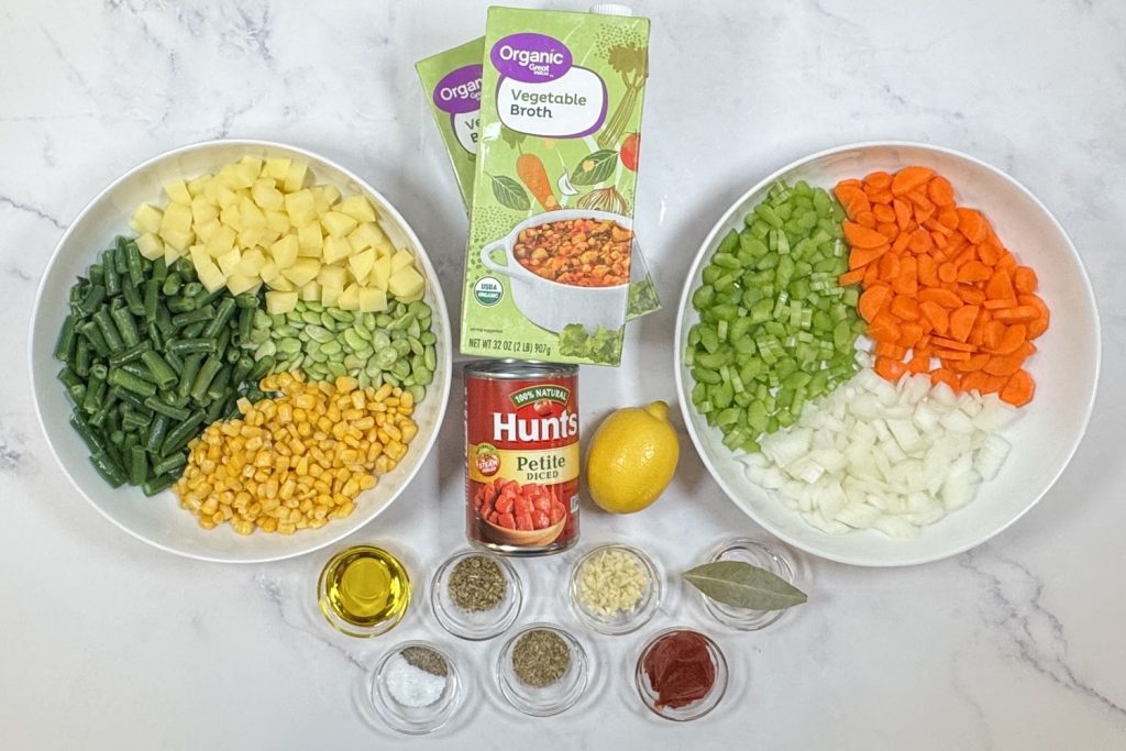 Ingredients for vegetable soup arranged on a marble surface, including diced potatoes, green beans, lima beans, corn, carrots, celery, onion, vegetable broth, petite diced tomatoes, lemon, olive oil, garlic, tomato paste, bay leaf, and dried herbs.