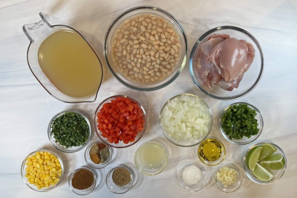 Ingredients for white bean chicken chili including chicken thighs, white beans, chicken broth, corn, diced red and green bell peppers, onion, garlic, lime wedges, olive oil, chopped cilantro, and chili spices.