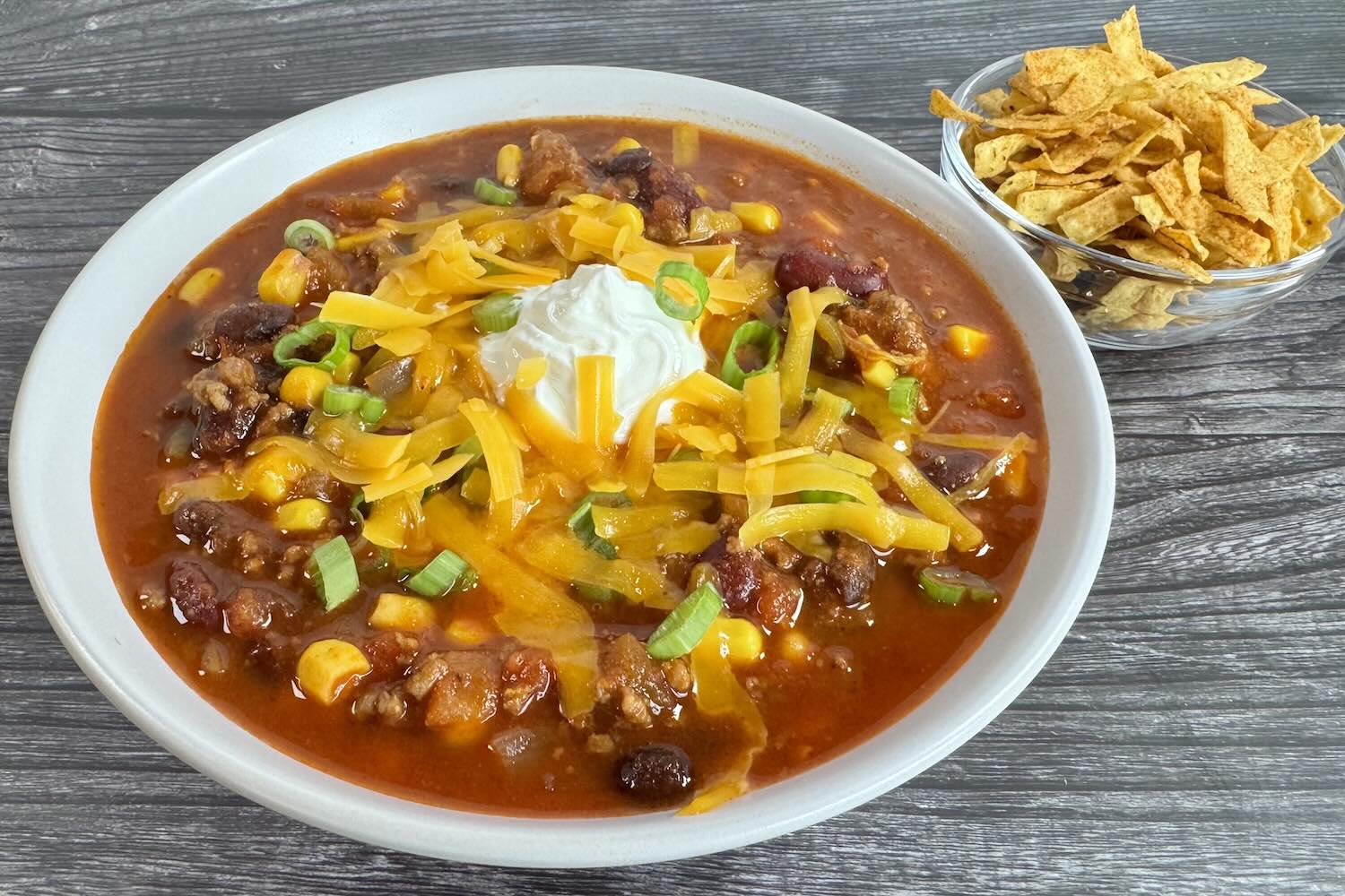 Bowl of easy taco soup topped with shredded cheddar cheese, sour cream, and sliced green onions, served with tortilla chips on the side.