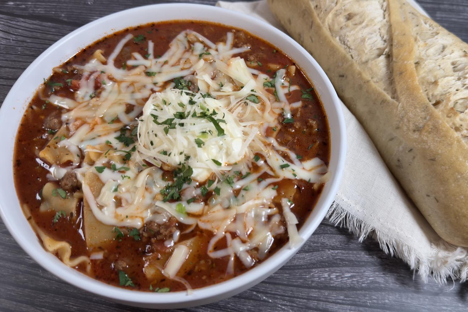 Bowl of lasagna soup topped with melted mozzarella, ricotta, and fresh herbs, served with crusty bread on the side.