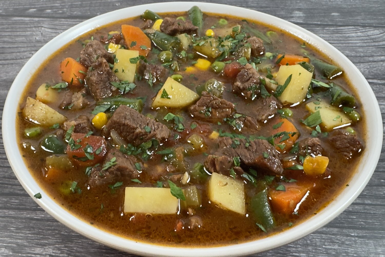 Bowl of beef vegetable soup with chunks of beef, potatoes, carrots, green beans, corn, and peas in a rich broth, garnished with fresh parsley.