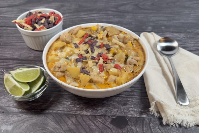 Bowl of chicken and poblano corn chowder topped with shredded cheese and tortilla strips, served with lime wedges on the side.