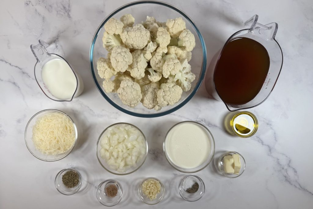 Ingredients for creamy cauliflower soup including cauliflower florets, broth, cream, onion, garlic, butter, olive oil, and Parmesan cheese.