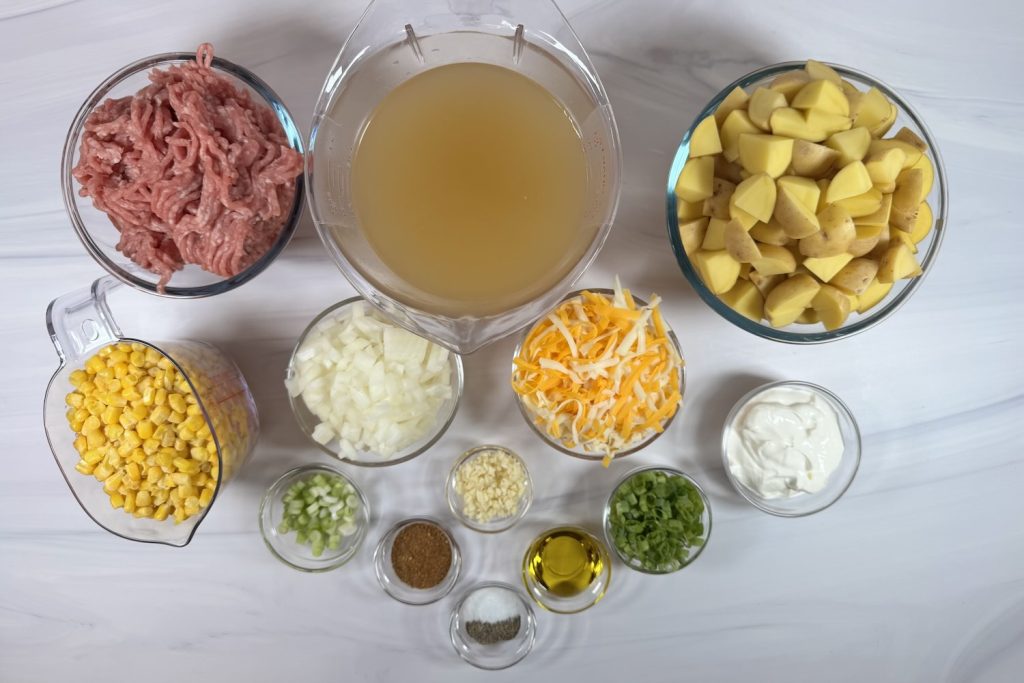 Ingredients for Santa Fe pork and potato soup including ground pork, potatoes, chicken broth, corn, onion, garlic, shredded cheese, sour cream, green onions, olive oil, taco seasoning, salt, and black pepper.