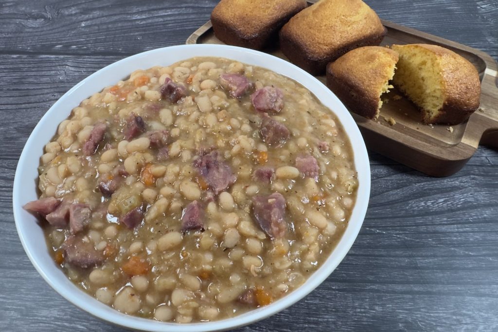 Bowl of ham and bean soup with tender white beans, chunks of ham, and vegetables, served with cornbread on the side.