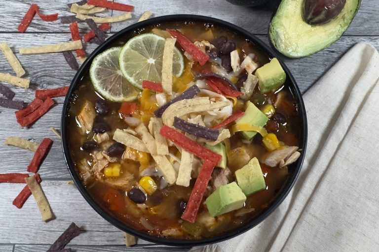 Bowl of chicken tortilla soup topped with tortilla strips, avocado, shredded cheese, and lime slices, with black beans, corn, and shredded chicken in a rich tomato broth.