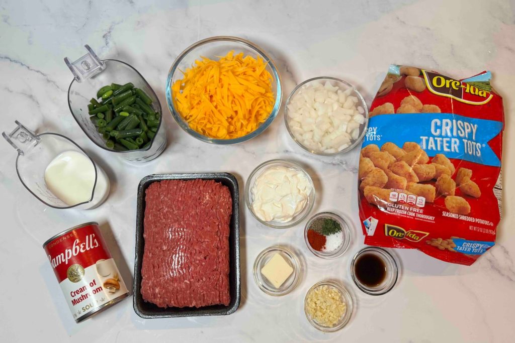 Ingredients for tater tot casserole including ground beef, frozen tater tots, green beans, cheddar cheese, onion, cream of mushroom soup, sour cream, milk, garlic, butter, Worcestershire sauce, and seasonings.