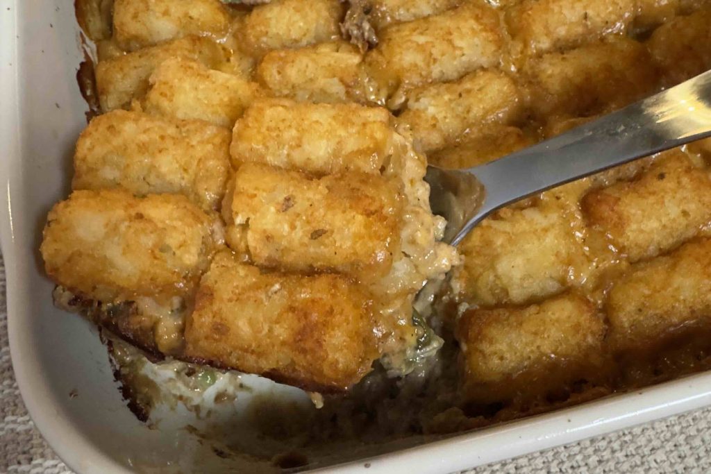Tater tot casserole being served from a white baking dish, showing crispy golden tater tots over a creamy ground beef, green bean, and cheddar cheese filling.