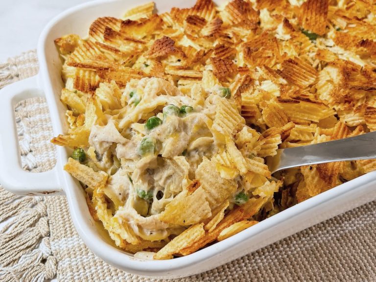 Tuna noodle casserole being scooped from a baking dish, showing creamy noodles, tuna, peas, melted cheese, and a golden potato chip topping.
