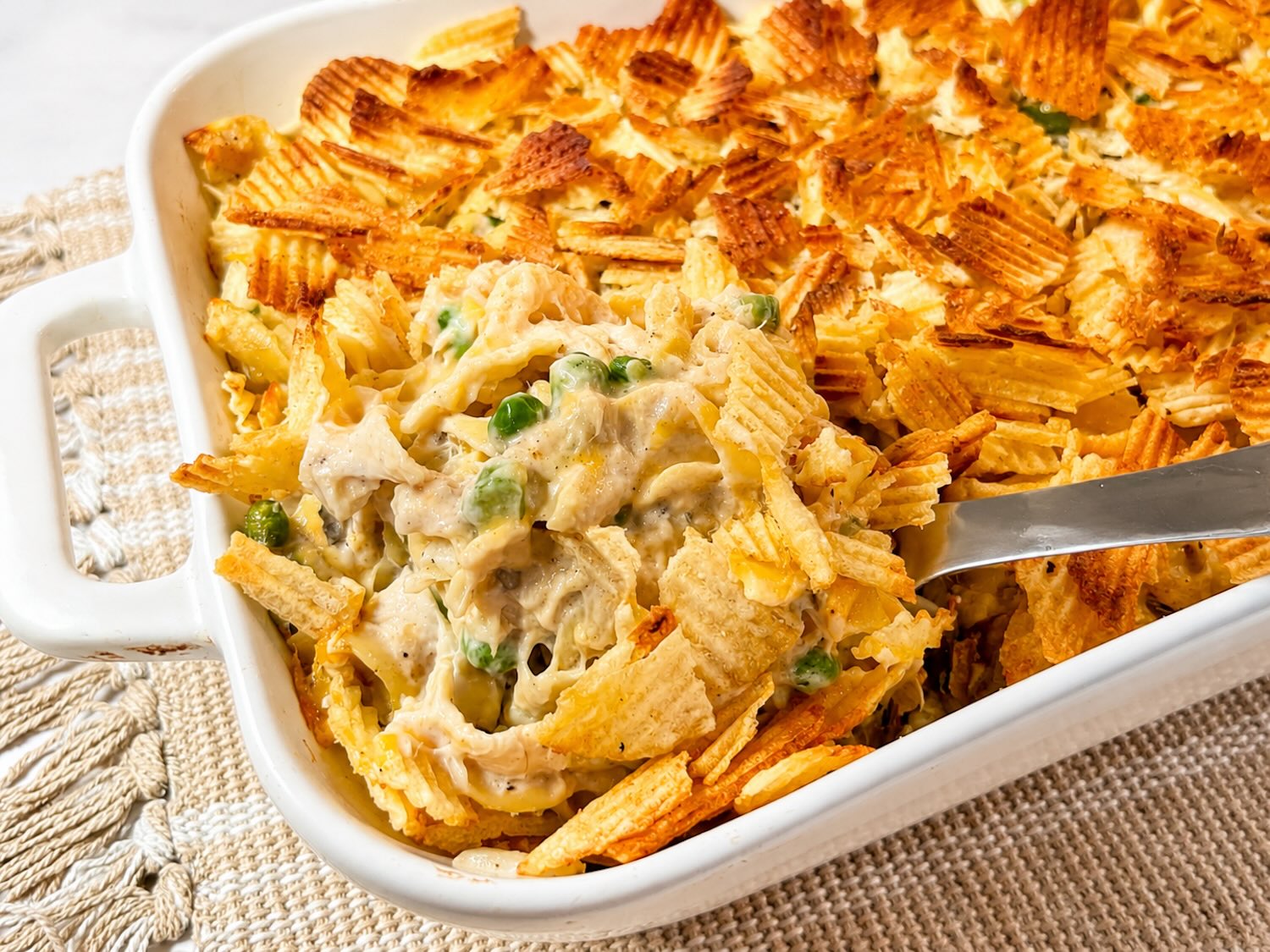 Tuna noodle casserole being scooped from a baking dish, showing creamy noodles, tuna, peas, melted cheese, and a golden potato chip topping.