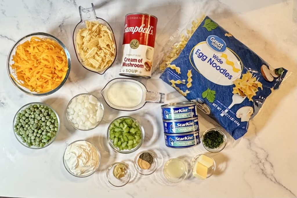 Ingredients for tuna noodle casserole including egg noodles, canned tuna, cream of mushroom soup, cheddar cheese, peas, onion, celery, sour cream, milk, potato chips, butter, lemon juice, Dijon mustard, parsley, and seasonings.