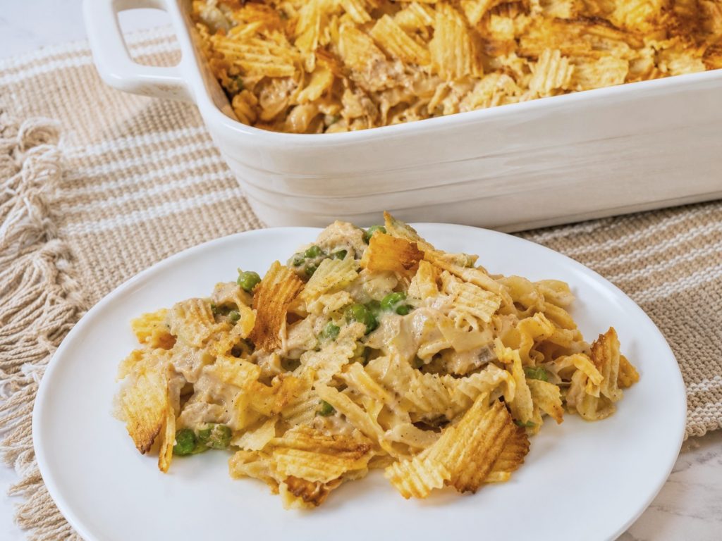 Plate of tuna noodle casserole with egg noodles, tuna, peas, cheddar cheese, and a crispy potato chip topping, served in front of the baked casserole dish.