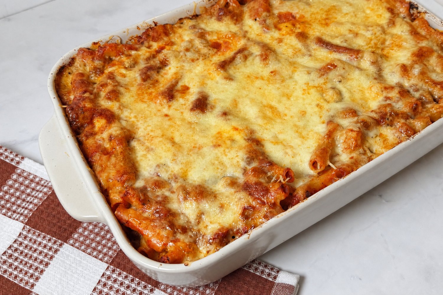 Baked ziti with ground beef in a white baking dish, topped with melted mozzarella and Parmesan cheese over ziti pasta and tomato meat sauce.
