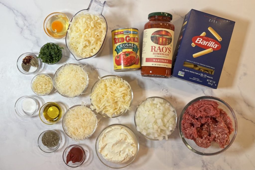 Ingredients for baked ziti with ground beef including ziti pasta, ground beef, marinara sauce, crushed tomatoes, ricotta, mozzarella, Parmesan, onion, garlic, egg, olive oil, parsley, tomato paste, and seasonings.