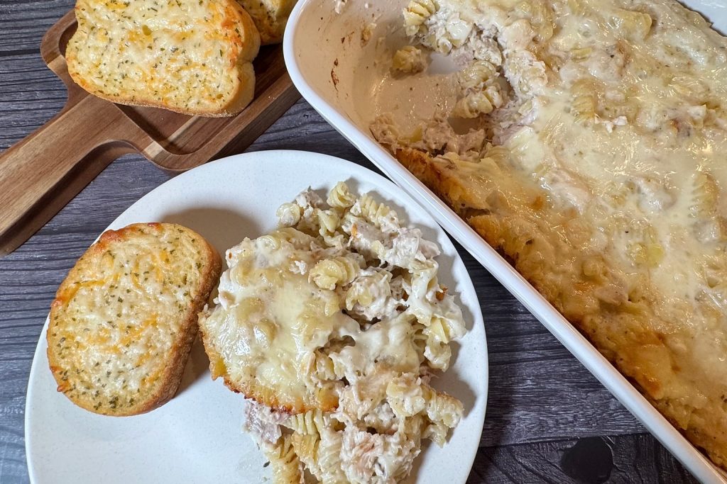 Serving of Chicken Alfredo Bake on a plate with garlic bread, showing rotini pasta, chicken, creamy Alfredo sauce, and melted mozzarella beside the casserole dish.
