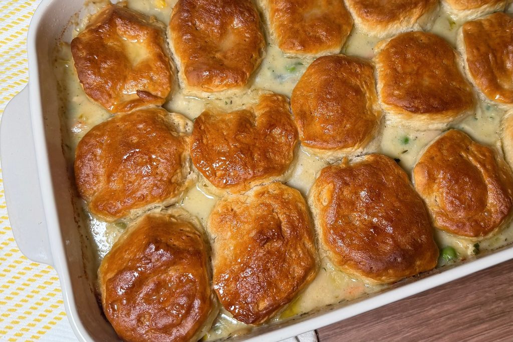 Chicken pot pie casserole baked in a white dish with golden buttermilk biscuits over a creamy chicken, pea, carrot, celery, and corn filling.