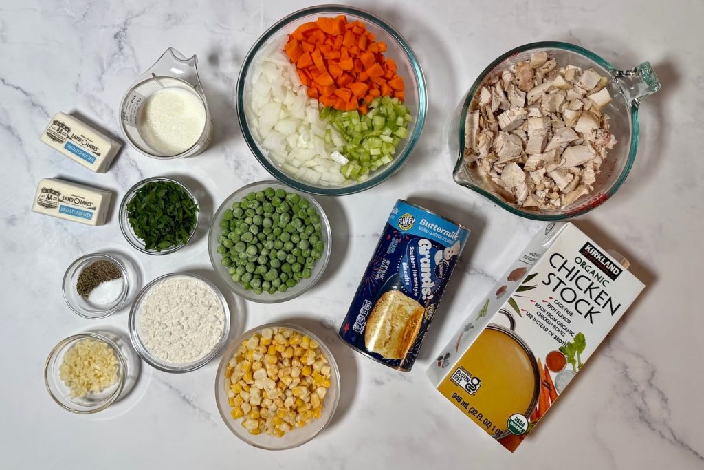 Ingredients for chicken pot pie casserole including cooked chicken, buttermilk biscuits, chicken stock, milk, butter, flour, peas, corn, onion, carrots, celery, garlic, parsley, and seasonings.