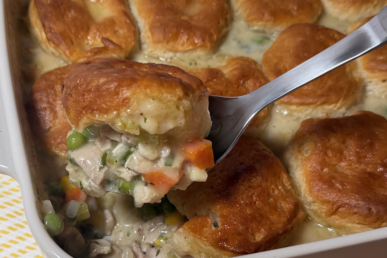 Chicken pot pie casserole being served from a white baking dish, showing a golden biscuit topping over creamy chicken, peas, carrots, celery, and corn filling.
