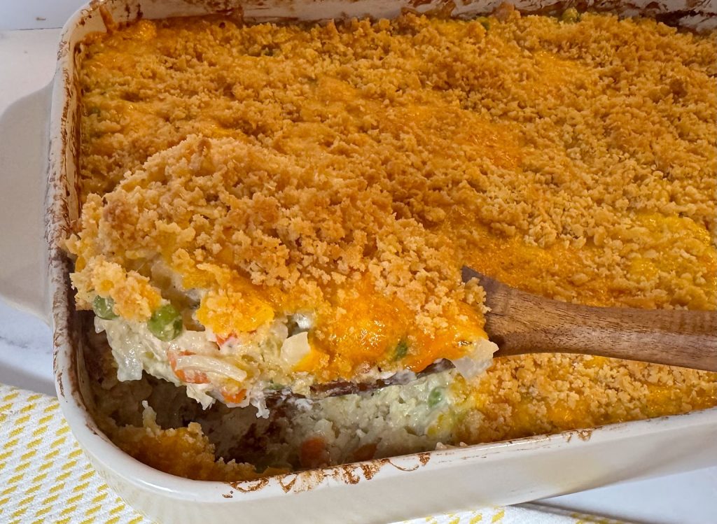 Creamy chicken and rice casserole being served from a white baking dish, with chicken, rice, peas, carrots, cheddar cheese, and a buttery cracker topping.