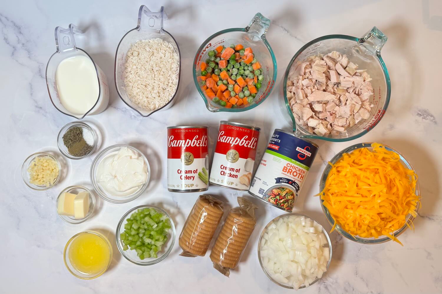 Ingredients for creamy chicken and rice casserole including cooked chicken, rice, mixed vegetables, cheddar cheese, cream of chicken soup, cream of celery soup, chicken broth, milk, onion, celery, butter, crackers, garlic, and seasonings.