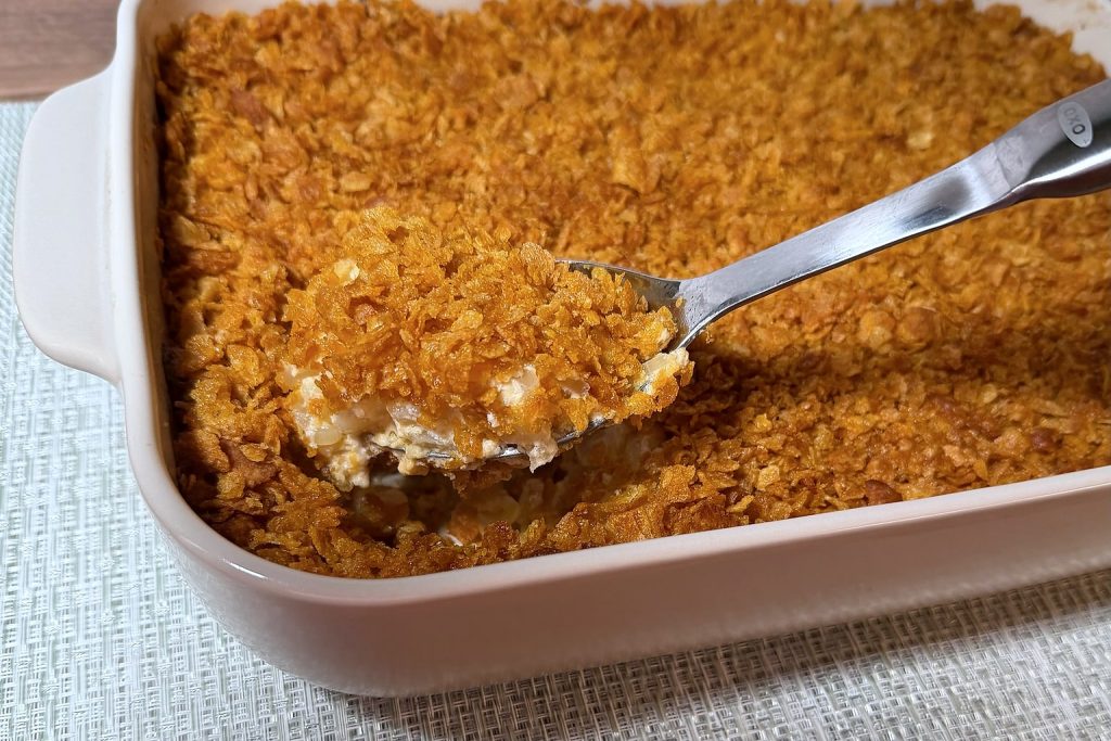 Funeral potatoes being served from a white baking dish, with a creamy hash brown potato filling under a golden, crunchy cornflake topping.