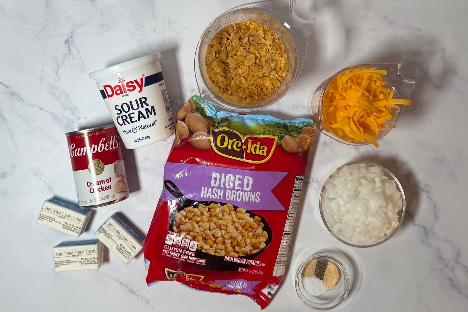 Ingredients for funeral potatoes including diced hash browns, cream of chicken soup, sour cream, cheddar cheese, onion, butter, cornflakes, garlic powder, onion powder, salt, and black pepper.