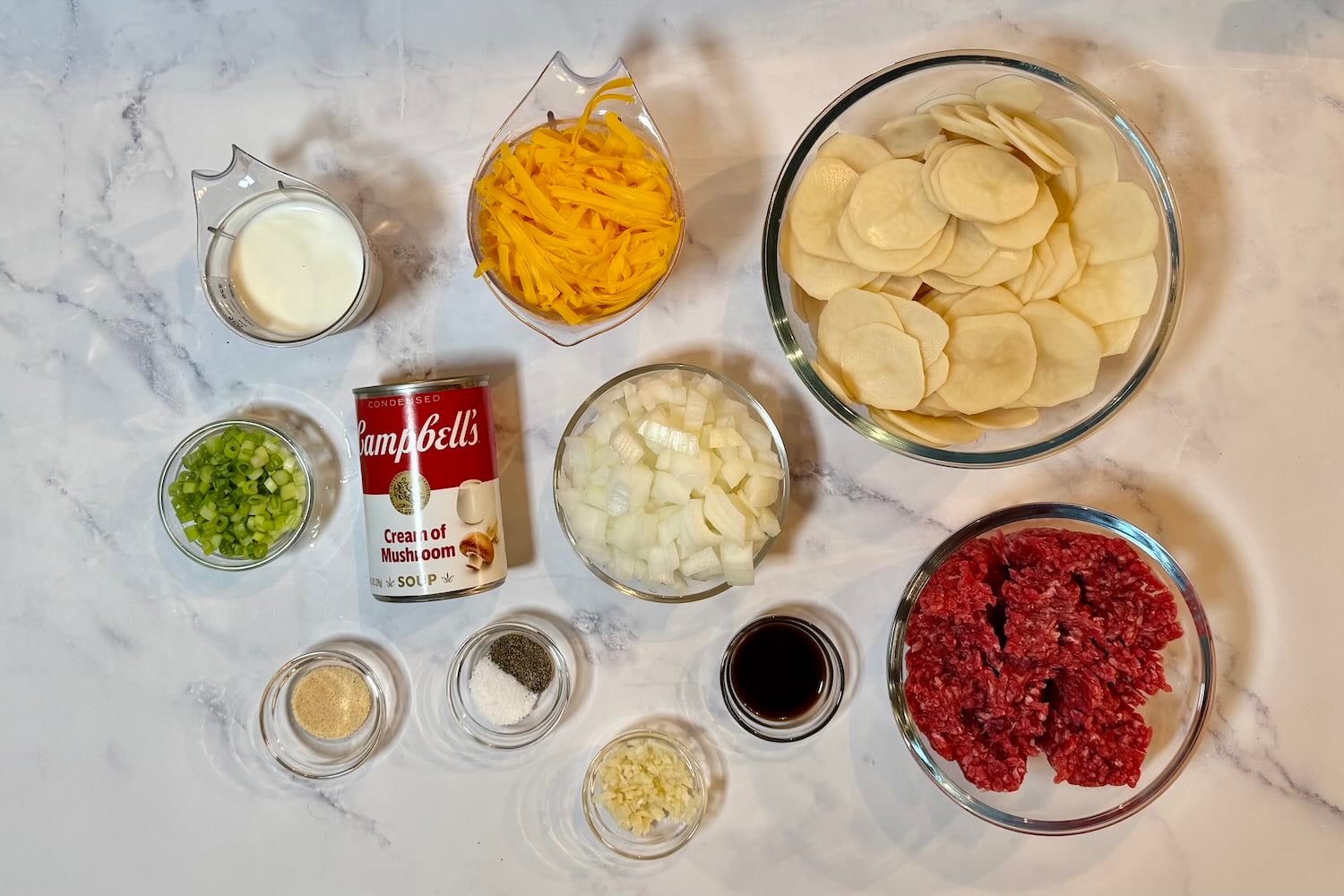 Ingredients for Hamburger Potato Casserole including ground beef, sliced potatoes, cheddar cheese, cream of mushroom soup, milk, onion, green onions, garlic, Worcestershire sauce, salt, pepper, and onion powder.