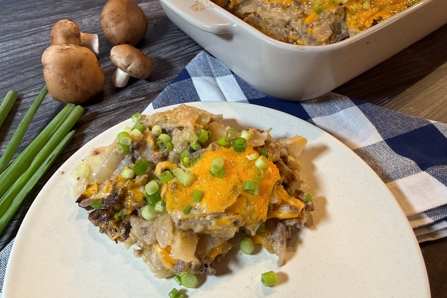 Serving of Hamburger Potato Casserole on a plate, showing layers of ground beef, sliced potatoes, creamy mushroom sauce, melted cheddar cheese, and green onions.
